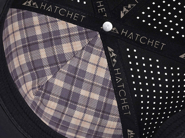 Close-up image of the interior of the Black colored Rover premium performance fitted hat from Hatchet Headwear. The interior of the hat features a black and tan plaid front panel liner, black seam tape with a taupe version of the Hatchet Headwear logo, and black sweatband featuring flex tech. Also visible in the image are diamond shaped laser perforations on the crown of the hat to provide breathability and reduce heat retention.