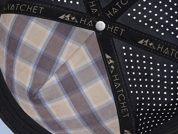 Close-up image of the interior of the Glacier colored Rover premium performance fitted hat from Hatchet Headwear. The interior of the hat features a tan, blue, and gray plaid front panel liner, black seam tape with a taupe version of the Hatchet Headwear logo, and black sweatband featuring flex tech. Also visible in the image are diamond shaped laser perforations on the crown of the hat to provide breathability and reduce heat retention.