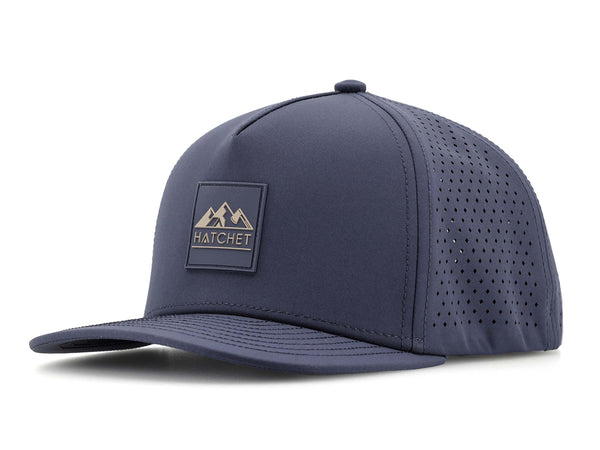 Slate Blue colored Rover premium performance fitted hat from Hatchet Headwear pictured with a white foreground and background. Hat features a color matched Slate Blue rubber logo patch with the Hatchet Headwear logo (mountains, tent, and hatchet above the word "HATCHET) in taupe. The hat is positioned at an approximately 45 degree angle to showcase the front and left-side profile of the hat.