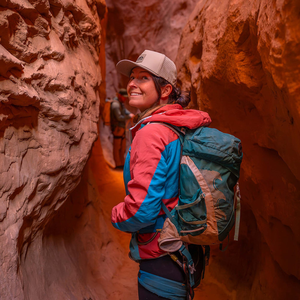 Woman wearing a Boulder colored Rover premium performance fitted hat from Hatchet Headwear, teal hiking backpack, red and blue hooded jacket, and outdoor gear while hiking through a narrow slot canyon with red rock walls.  In the blurred background, a man wearing outdoor clothing/gear and a Hunter Green colored Rover premium performance fitted hat from Hatchet Headwear is visible.