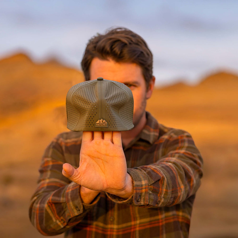 Man stretching a Hunter Green colored Rover fitted hat from Hatchet Headwear, to demonstrate the hat's adaptive flex tech properties, while wearing an army green, tan, and orange plaid shirt in an outdoor desert environment.  Background of the outdoor desert environment is blurred.