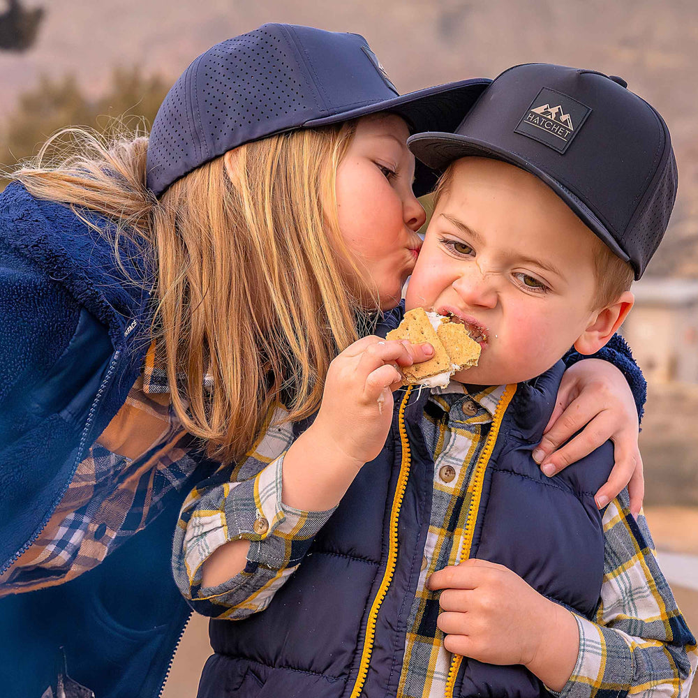 Young brother and sister wearing a Rover premium performance fitted hats from Hatchet Headwear.  The boy is wearing a Black colored Rover hat, a blue vest, and a blue, white and yellow plaid shirt while eating a s'more.  The sister is wearing a Slate Blue colored Rover hat, a blue fleece jacket, and a yellow, blue and white plaid shirt while kissing her younger brother on the cheek.  The brother and sister are in outdoors with a blurred desert landscape in the background.