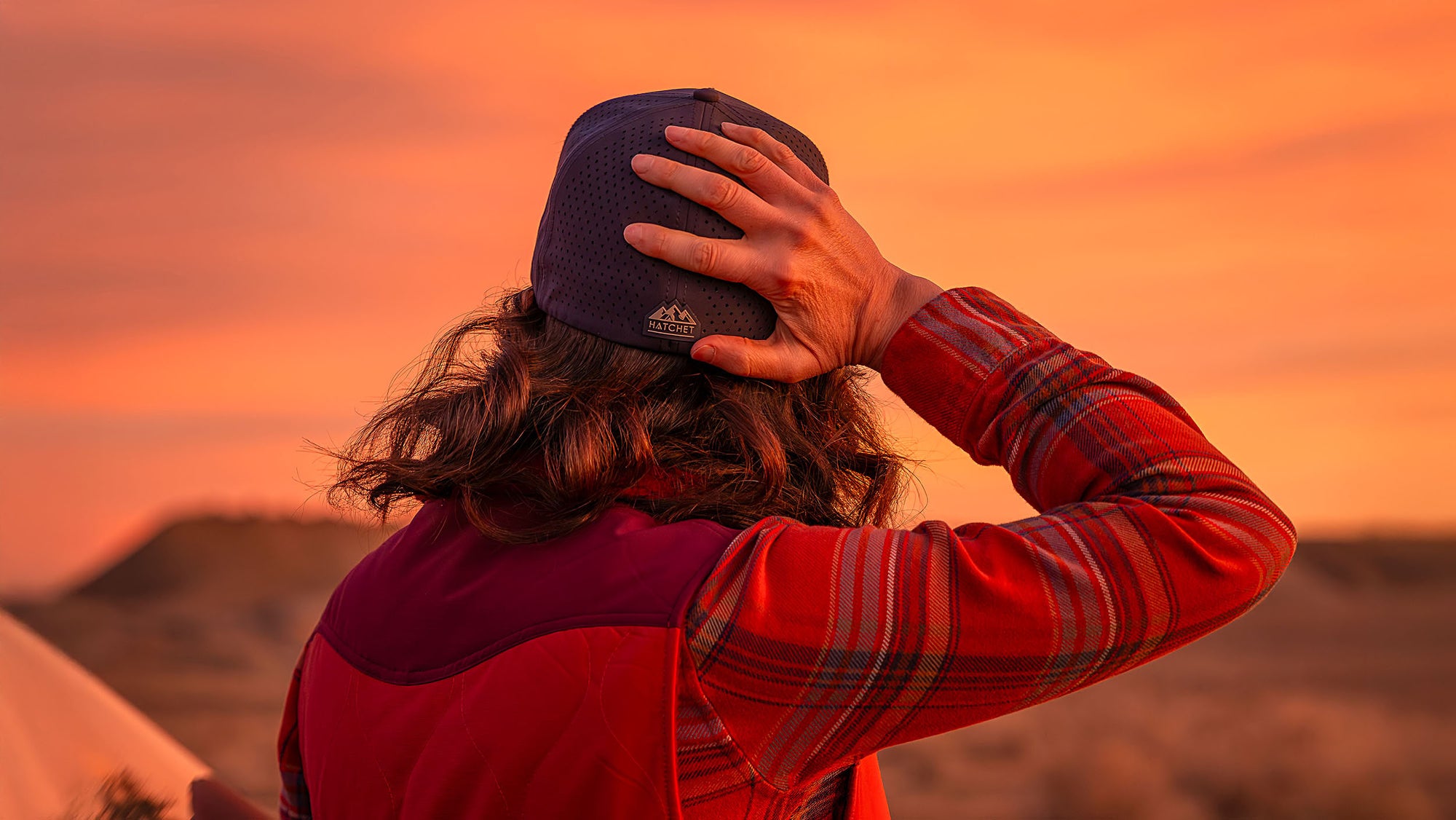 Woman wearing a Slate Blue colored Rover premium performance fitted hat from Hatchet Headwear, an orange plaid shirt, and orange vest while leaning/resting against an SUV with a warm desert sunset in the background.  Woman is facing away from the camera and looking off into the distant desert while holding the back of her fitted hat.