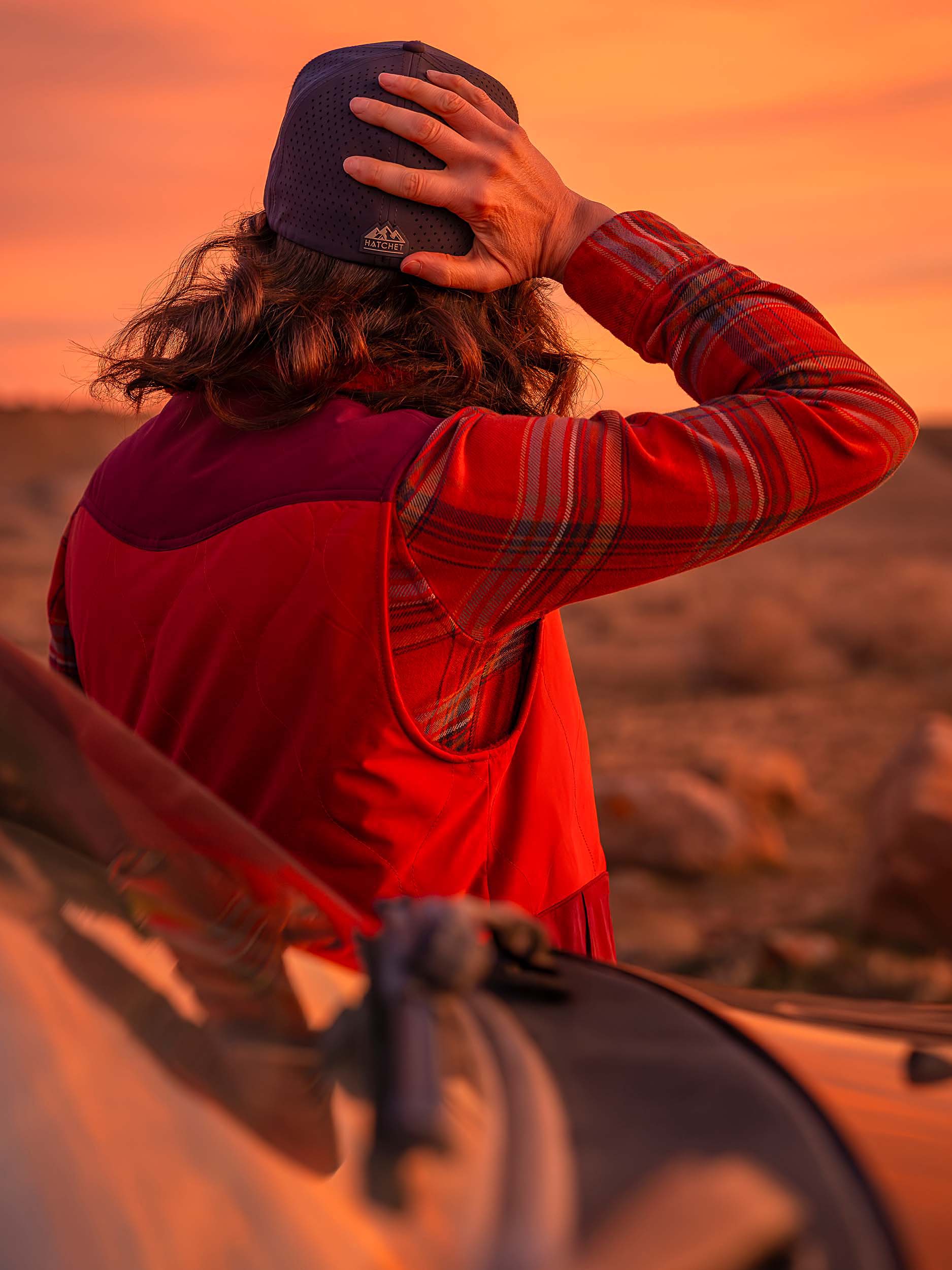 Woman wearing a Slate Blue colored Rover premium performance fitted hat from Hatchet Headwear, an orange plaid shirt, and orange vest while leaning/resting against an SUV with a warm desert sunset in the background.  Woman is facing away from the camera and looking off into the distant desert while holding the back of her fitted hat.