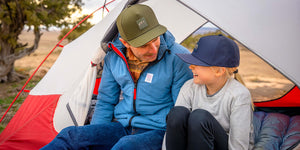 Father and daughter wearing Rover premium performance fitted hats from Hatchet Headwear while sitting at the entrance of a tent with a high desert landscape in the background. Father is wearing a Hunter Green colored Rover, a blue jacket, a brown plaid shirt and blue jeans. Daughter is smiling and wearing a Steel Blue Rover, gray long-sleeve shirt, and black leggings.