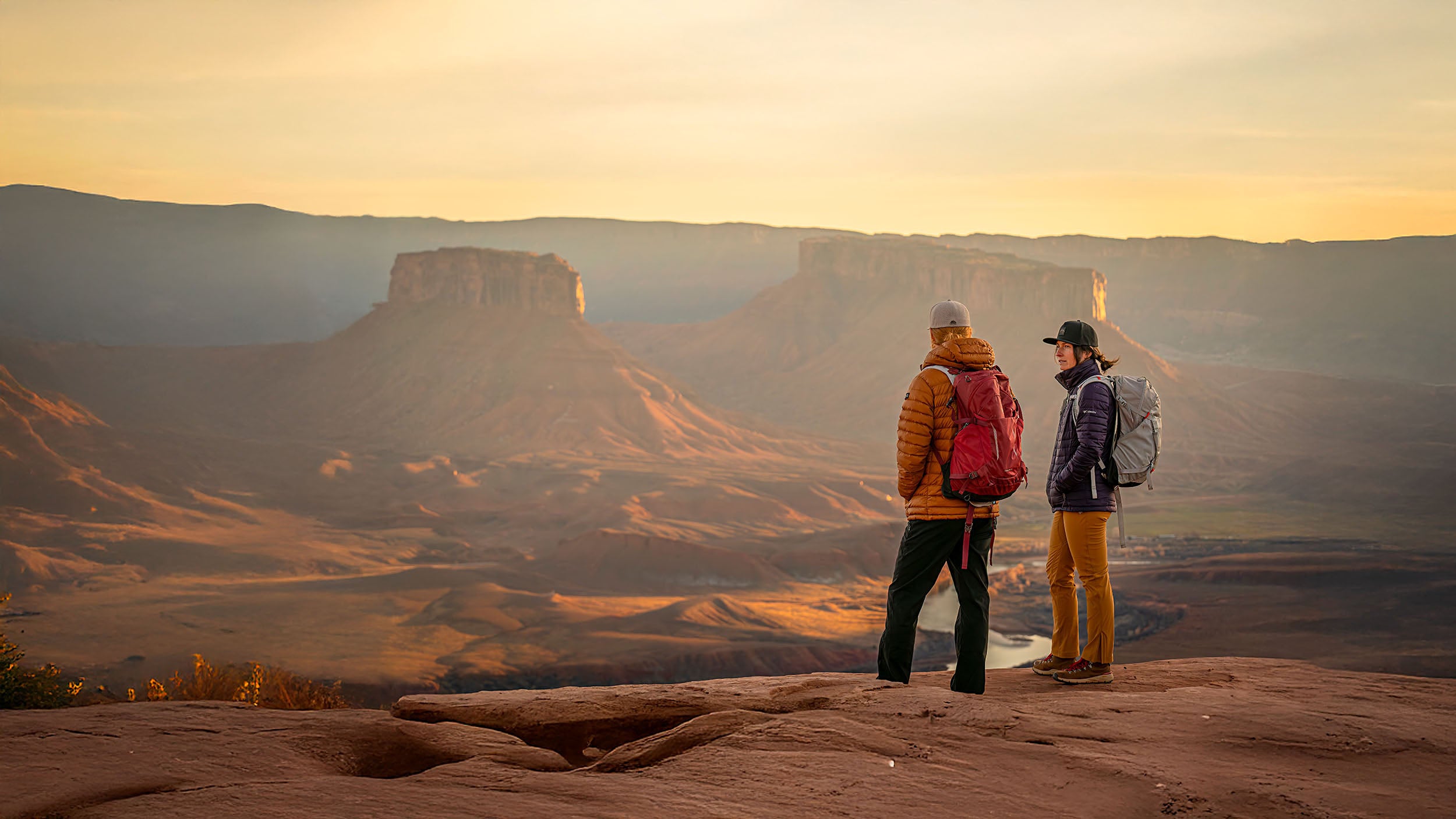 A man and a woman standing on a rocky outcrop overlooking a red rock canyon at sunset.  The man is wearing a Boulder colored Rover premium performance fitted from Hatchet Headwear, a burnt orange hooded jacket, dark pants, and a red backpack.  The woman is wearing a Black colored premium performance fitted hat from Hatchet Headwear, a purple jacket, orange pants, and a light gray backpack.