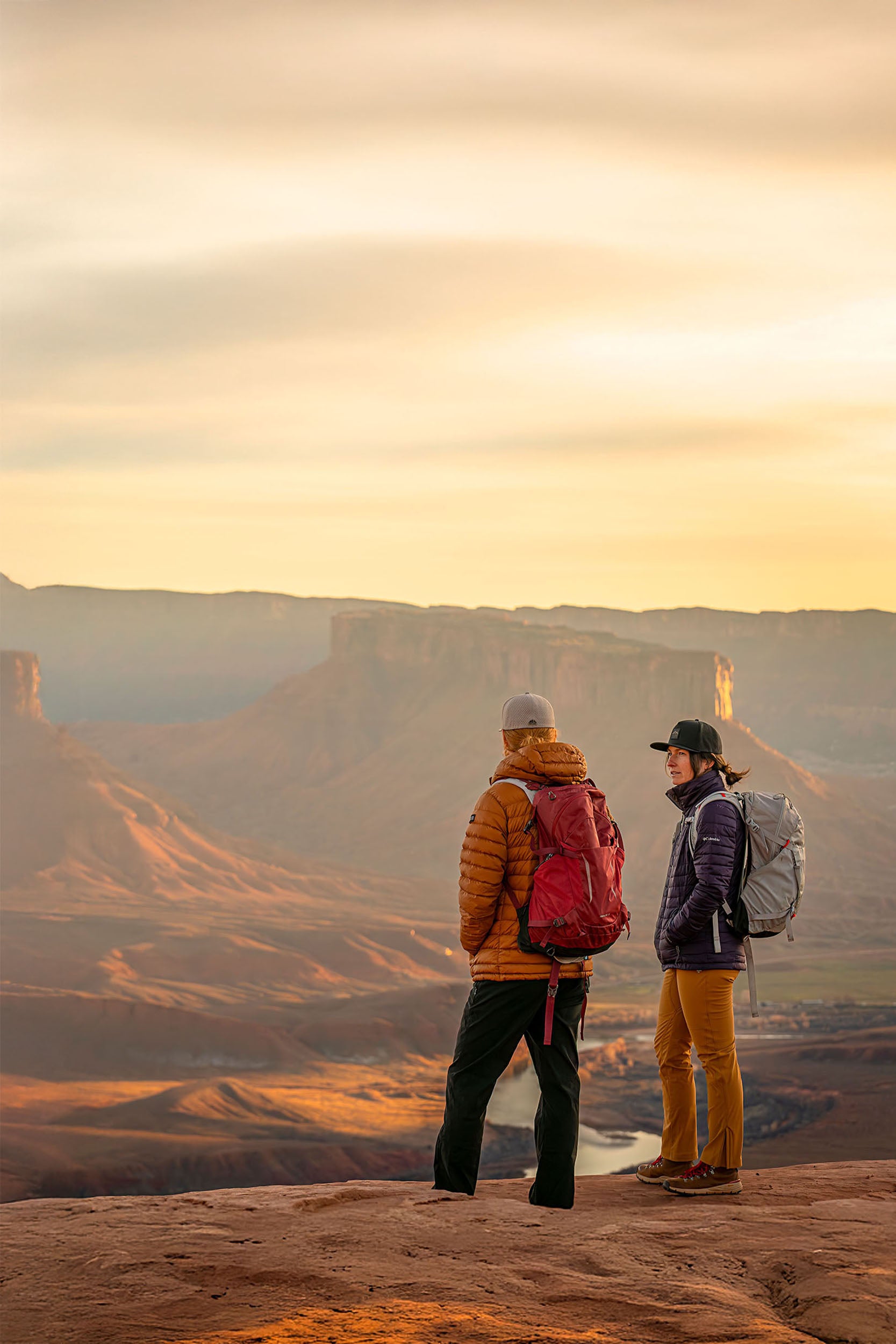 A man and a woman standing on a rocky outcrop overlooking a red rock canyon at sunset.  The man is wearing a Boulder colored Rover premium performance fitted from Hatchet Headwear, a burnt orange hooded jacket, dark pants, and a red backpack.  The woman is wearing a Black colored premium performance fitted hat from Hatchet Headwear, a purple jacket, orange pants, and a light gray backpack.