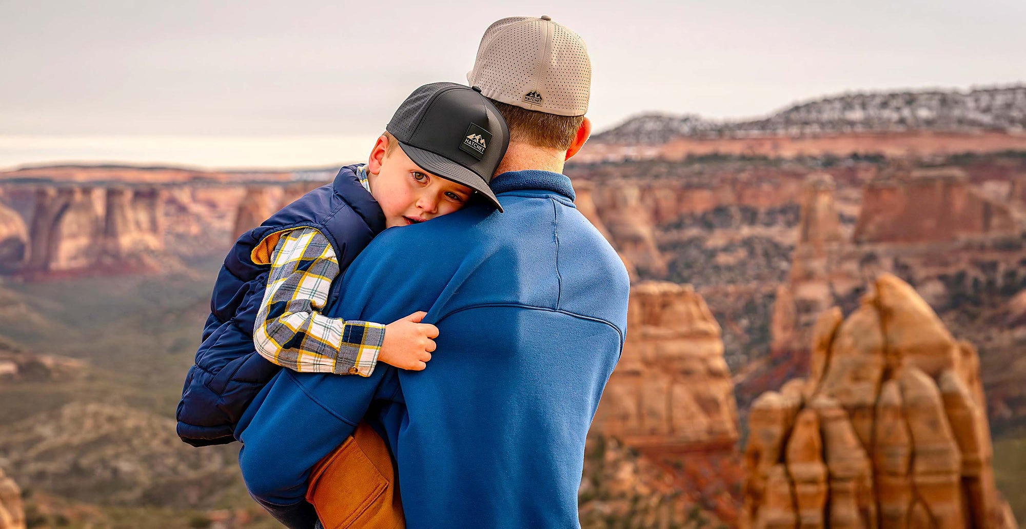 Father and son wearing Rover premium performance fitted hats from Hatchet Headwear. Father is wearing a Boulder colored Rover hat and blue jacket while holding his son in his arms and looking away from the camera at a desert canyon view.  Son is looking at the camera and wearing a Black colored Rover hat, Blue vest, brown cargo pants, and a blue, white and yellow plaid shirt.
