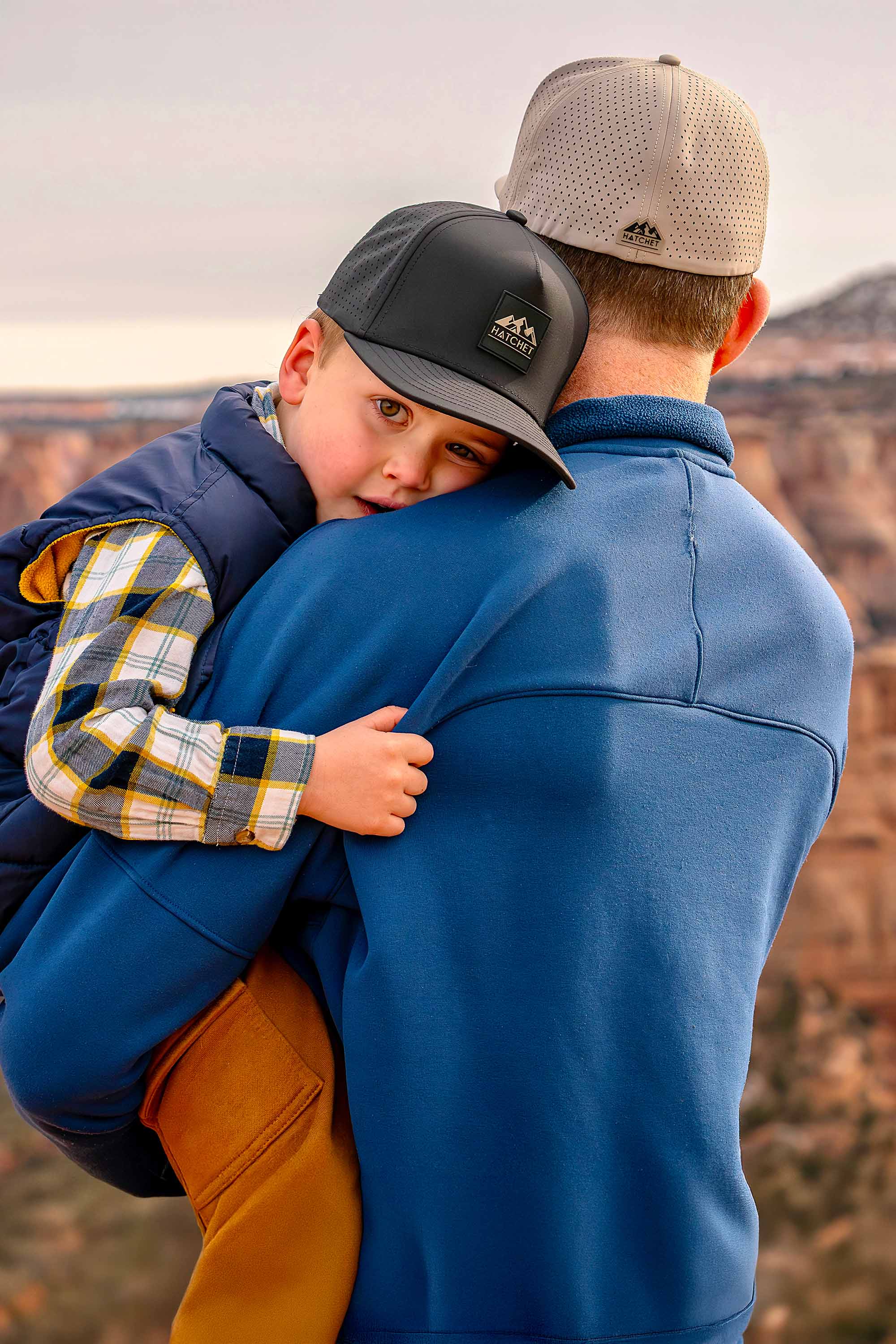 Father and son wearing Rover premium performance fitted hats from Hatchet Headwear. Father is wearing a Boulder colored Rover hat and blue jacket while holding his son in his arms and looking away from the camera at a desert canyon view.  Son is looking at the camera and wearing a Black colored Rover hat, Blue vest, brown cargo pants, and a blue, white and yellow plaid shirt.