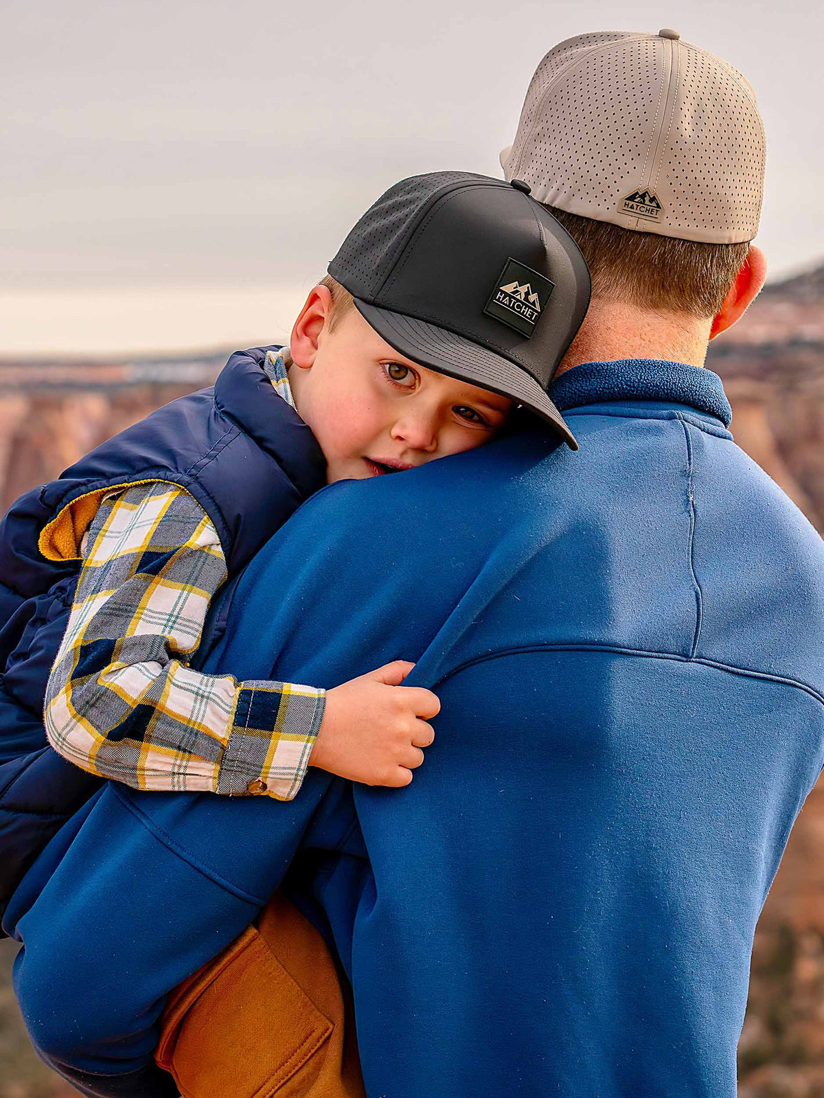 Father and son wearing Rover premium performance fitted hats from Hatchet Headwear. Father is wearing a Boulder colored Rover hat and blue jacket while holding his son in his arms and looking away from the camera at a desert canyon view.  Son is looking at the camera and wearing a Black colored Rover hat, Blue vest, brown cargo pants, and a blue, white and yellow plaid shirt.