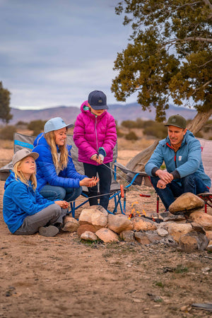 A Father and Mother are sitting fireside at a campsite with their young son and young daughter while roasting hot dogs over an open campfire flame. Each of the family members is wearing a different colored Rover premium performance fitted hat from Hatchet Headwear and outdoor styled clothing. A blurred desert landscape is in the background.