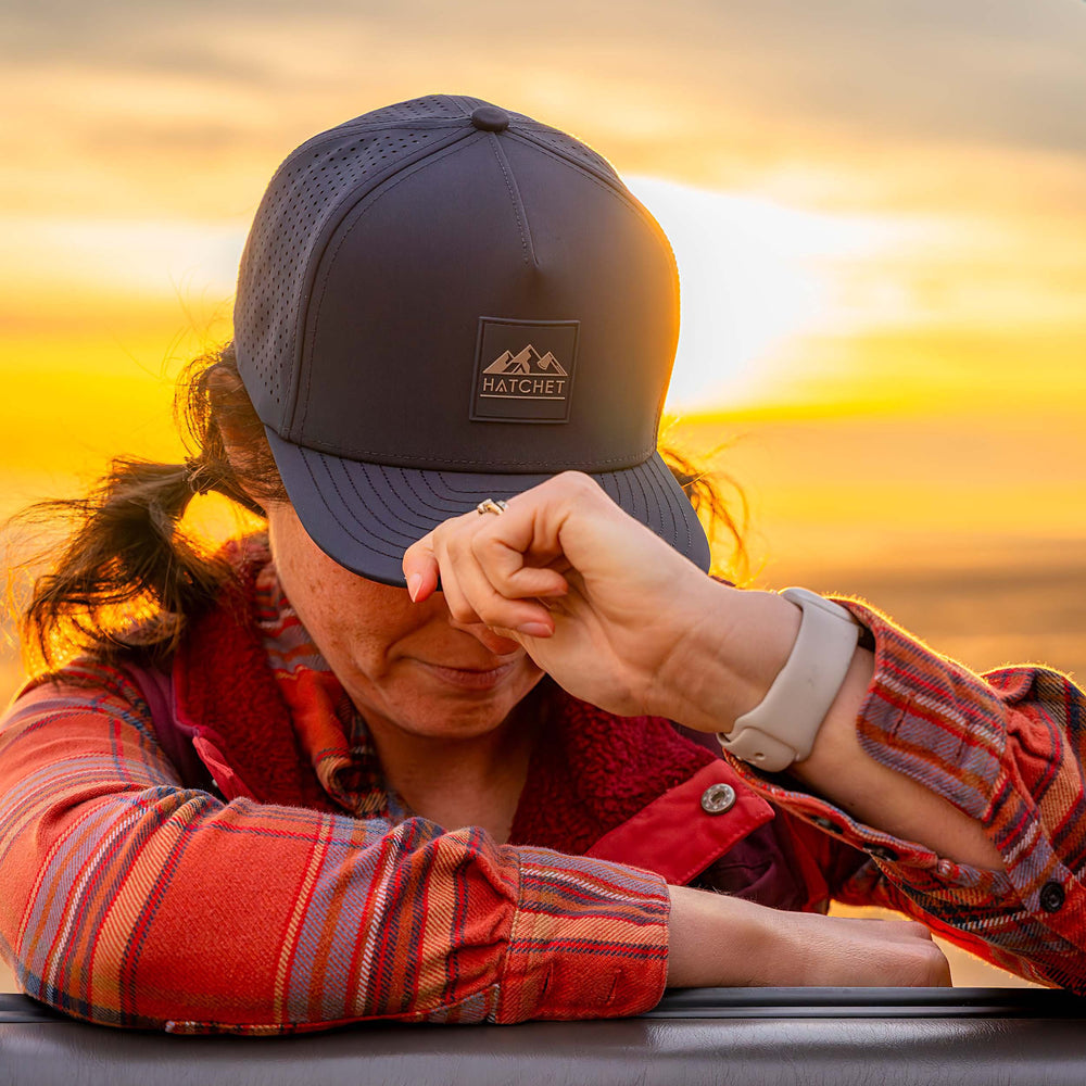 Woman wearing a Slate Blue colored Rover premium performance fitted hat from Hatchet Headwear, an orange plaid shirt, and orange vest while leaning/resting on the open window of vehicle with a warm sunset in the background.