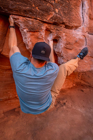 Man wearing a Black colored Rover premium performance fitted hat from Hatchet Headwear, blue short-sleeved shirt, and tan pants while climbing a red rock wall.