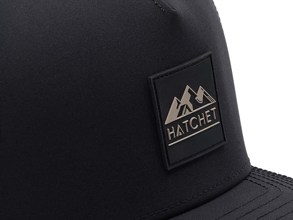 Close-up image of the Black colored Rover premium performance hat from Hatchet Headwear.  The image depicts the Hatchet Headwear rubber patch logo on the front of the hat in detail.  The taupe and Black colored logo features mountains, a tent, and a hatchet above the word "HATCHET".  The hat is positioned in front of a white background.
