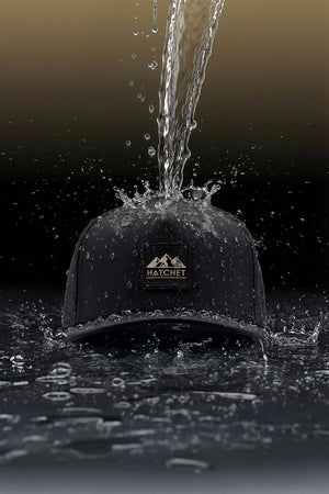 Black colored Rover premium performance fitted hat from Hatchet Headwear being splashed by water, to demonstrate the hat's water repellent properties, in front of a dark gradient background.