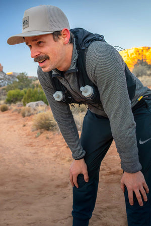 Man wearing a Boulder colored Rover premium performance fitted hat from Hatchet Headwear, long-sleeved gray waffle shirt, black trail running pants, and hydration vest while taking a break from running to catch his breath in an outdoor desert environment.