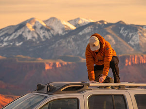 A man crouched on the roof of a light gray SUV that is parked in front of  a red rock canyon and snow covered mountains at sunset.  The man is wearing a Boulder colored Rover premium performance fitted hat from Hatchet Headwear, a burnt orange hooded jacket, and dark hiking pants.