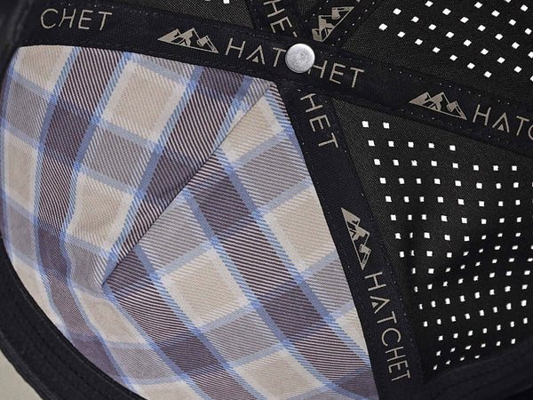 Close-up image of the interior of the Boulder colored Rover premium performance fitted hat from Hatchet Headwear.  The interior of the hat features a tan, blue, and gray plaid front panel liner, black seam tape with a taupe version of the Hatchet Headwear logo, and black sweatband featuring flex tech.  Also visible in the image are diamond shaped laser perforations on the crown of the hat to provide breathability and reduce heat retention.
