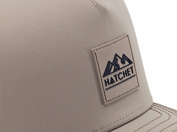 Close-up image of the Boulder colored Rover premium performance hat from Hatchet Headwear.  The image depicts the Hatchet Headwear rubber patch logo on the front of the hat in detail.  The black and boulder colored logo features mountains, a tent, and a hatchet above the word "HATCHET".  The hat is positioned in front of a white background.