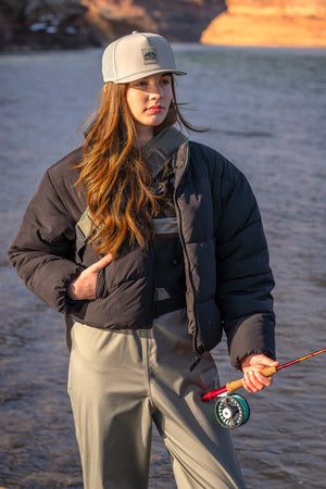 Woman wearing a Boulder colored Rover premium performance fitted hat from Hatchet Headwear, fly fishing bibs, and a black puffer jacket while standing in a lake and holding a fly fishing rod.