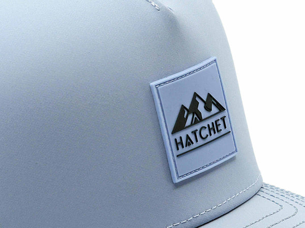 Close-up image of the Glacier colored Rover premium performance hat from Hatchet Headwear.  The image depicts the Hatchet Headwear rubber patch logo on the front of the hat in detail.  The Slate Blue and  Glacier colored logo features mountains, a tent, and a hatchet above the word "HATCHET".  The hat is positioned in front of a white background.