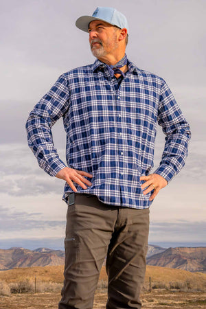 Man wearing a Glacier colored Rover premium performance fitted hat from Hatchet Headwear, blue plaid shirt, and gray hiking pants while looking into the distance of an outdoor desert environment. 