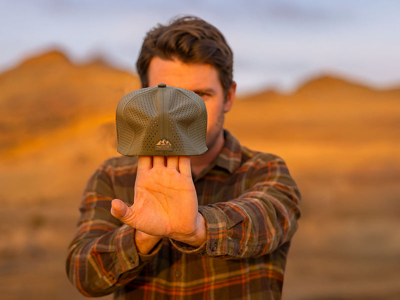 Man stretching a Hunter Green colored Rover fitted hat from Hatchet Headwear, to demonstrate the hat's adaptive flex tech properties, while wearing an army green, tan, and orange plaid shirt in an outdoor desert environment.  Background of the outdoor desert environment is blurred.