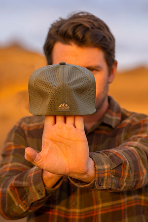 Man stretching a Hunter Green colored Rover fitted hat from Hatchet Headwear, to demonstrate the hat's adaptive flex tech properties, while wearing an army green, tan, and orange plaid shirt in an outdoor desert environment.  Background of the outdoor desert environment is blurred.