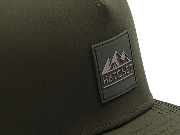 Close-up image of the Hunter Green colored Rover premium performance hat from Hatchet Headwear.  The image depicts the Hatchet Headwear rubber patch logo on the front of the hat in detail.  The taupe and Hunter Green colored logo features mountains, a tent, and a hatchet above the word "HATCHET".  The hat is positioned in front of a white background.