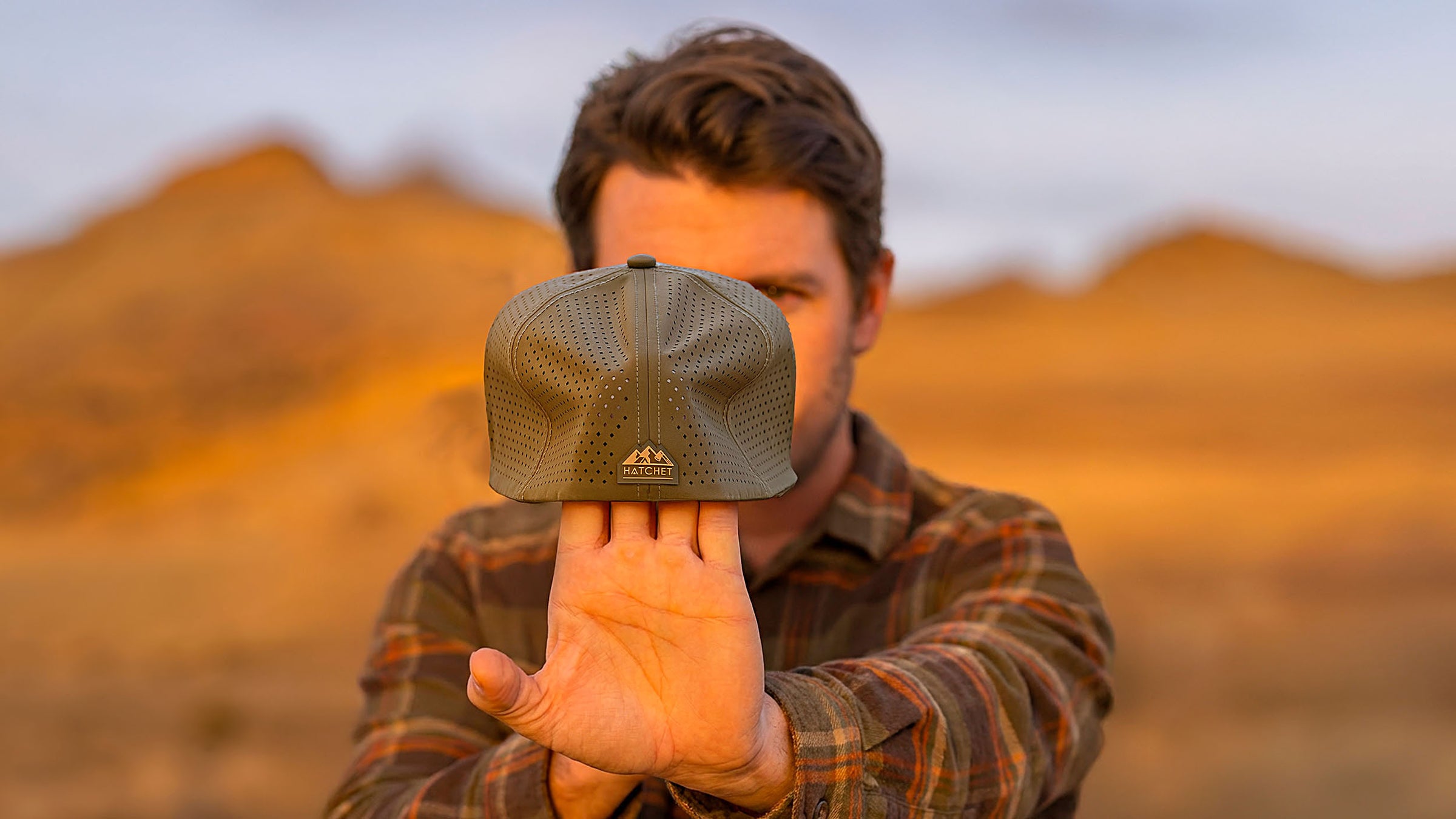 Man stretching a Hunter Green colored Rover premium performance fitted hat from Hatchet Headwear, to demonstrate the hat's adaptive flex tech properties, while wearing an army green, tan, and orange plaid shirt in an outdoor desert environment.  Background of the outdoor desert environment is blurred.