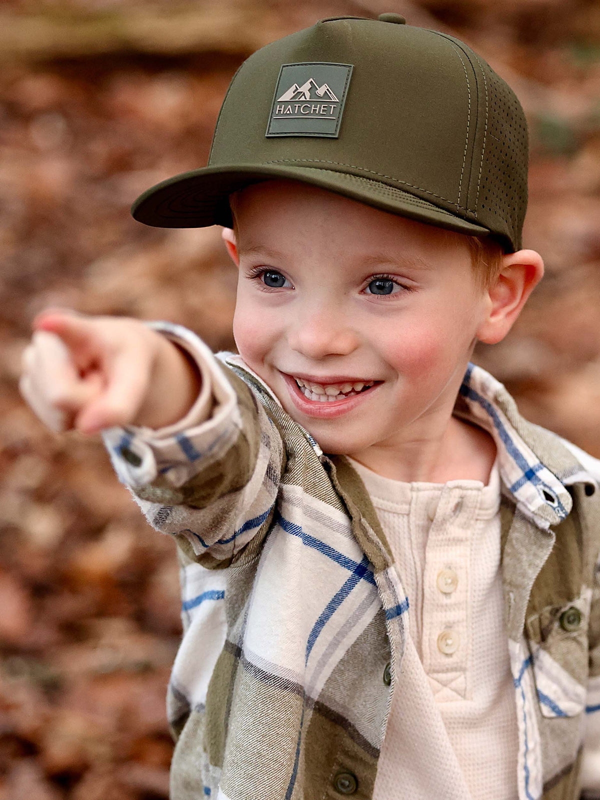 Young boy standing in a forest with autumn leaves in the blurred background.  The boy is smiling and pointing towards the horizon.  The boy is wearing a Hunter Green colored Rover premium performance fitted hat from Hatchet Headwear, unbuttoned green plaid shirt, and a cream waffled henley.