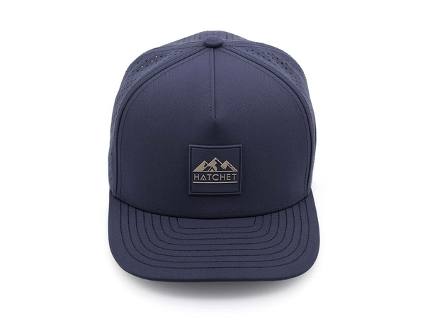 Slate Blue colored Rover premium performance fitted hat from Hatchet Headwear pictured with a white foreground and background.  Hat features a color matched Slate Blue rubber logo patch with the Hatchet Headwear logo (mountains, tent, and  hatchet above the word "HATCHET) in taupe.  The hat is pictured from the front and facing directly at the camera.  Camera position is elevated and front facing image of the hat is captured from above.