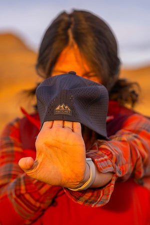 Female stretching a Slate Blue colored Rover premium performance fitted hat from Hatchet Headwear, to demonstrate the hat's adaptive flex tech properties, while wearing an orange plaid shirt and orange vest in an outdoor desert environment.  Background of the outdoor desert environment is blurred.