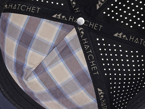 Close-up image of the interior of the Slate Blue colored Rover premium performance fitted hat from Hatchet Headwear.  The interior of the hat features a tan, blue, and gray plaid front panel liner, black seam tape with a taupe version of the Hatchet Headwear logo, and black sweatband featuring flex tech.  Also visible in the image are diamond shaped laser perforations on the crown of the hat to provide breathability and reduce heat retention.