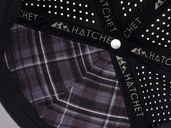 Close-up image of the interior of the Smoke colored Rover premium performance fitted hat from Hatchet Headwear.  The interior of the hat features a gray, black, and white plaid front panel liner, black seam tape with a taupe version of the Hatchet Headwear logo, and black sweatband featuring flex tech.  Also visible in the image are diamond shaped laser perforations on the crown of the hat to provide breathability and reduce heat retention.