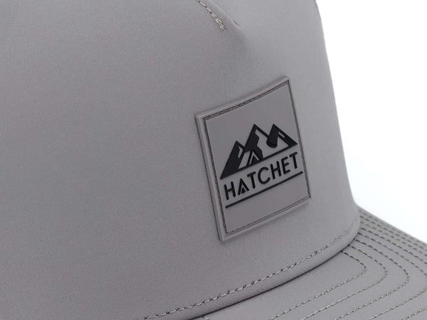 Close-up image of the Smoke colored Rover premium performance hat from Hatchet Headwear.  The image depicts the Hatchet Headwear rubber patch logo on the front of the hat in detail.  The Smoke and  black colored logo features mountains, a tent, and a hatchet above the word "HATCHET".  The hat is positioned in front of a white background.