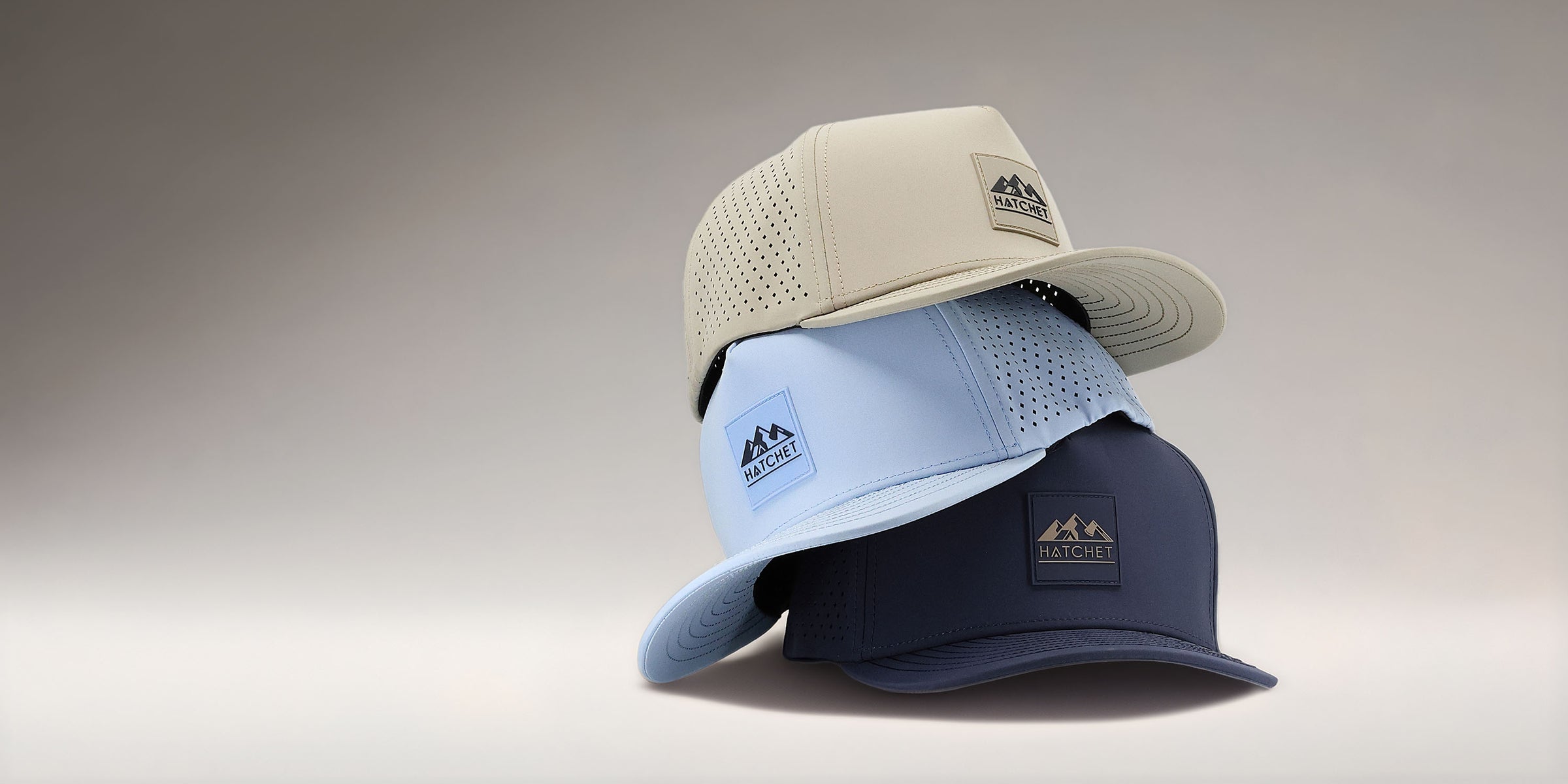 Three Rover premium performance fitted hats from Hatchet Headwear stacked on top of each other. The hats are sitting in front of a tan gradient background. From top to bottom, the featured hat colors are Boulder, Glacier, and Slate Blue.