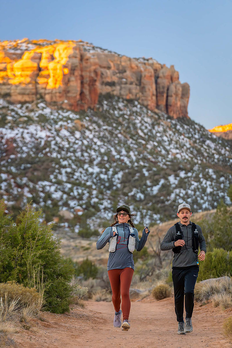 A man and woman running on a desert trail with red rock mountains in the background.  The man is wearing a Boulder colored Rover premium performance fitted hat from Hatchet Headwear, a gray waffled long-sleeved shirt, black trail running pants and a hydration vest.  The woman is wearing a Hunter Green colored Rover premium performance fitted hat from Hatchet Headwear, a grey long sleeved shirt, burnt orange leggings and a hydration vest.