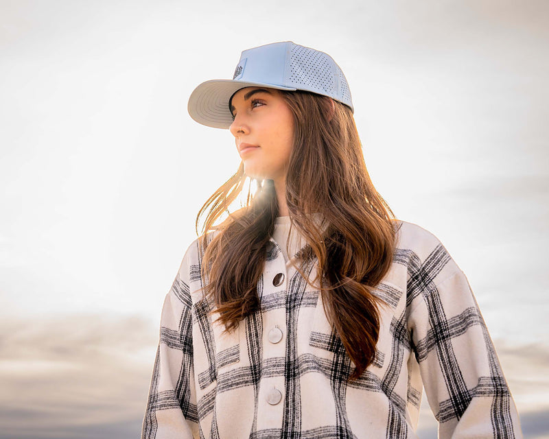 Woman wearing a Glacier colored Rover premium performance fitted hat from Hatchet Headwear and a black and white plaid shirt in front of a scenic sky with bright rays of sun.