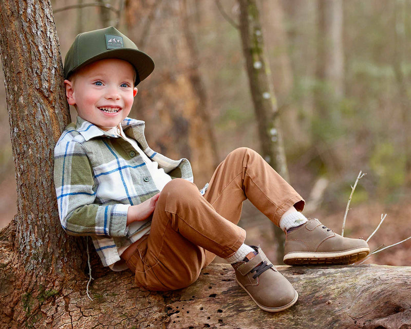 Young boy sitting on a log/leaning against a tree in a forest while wearing a Hunter Green colored Rover premium performance fitted hat from Hatchet Headwear, green plaid shirt, brown pants, and brown shoes.