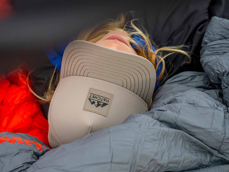 A person with long blonde hair wearing a Boulder colored Rover premium performance fitted hat from Hatchet Headwear and a black jacket with blue accents while sleeping in a tent on top of a gray and orange sleeping bag.