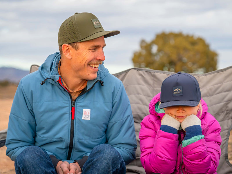 Father and daughter wearing Rover premium performance fitted hats from Hatchet Headwear while sitting in camping chairs with a high desert landscape in the background. Father is smiling while wearing a Hunter Green colored Rover hat, a blue jacket, a brown plaid shirt and blue jeans. Daughter has her chin resting on her fists while wearing a Steel Blue Rover hat and bright pink puffer jacket.