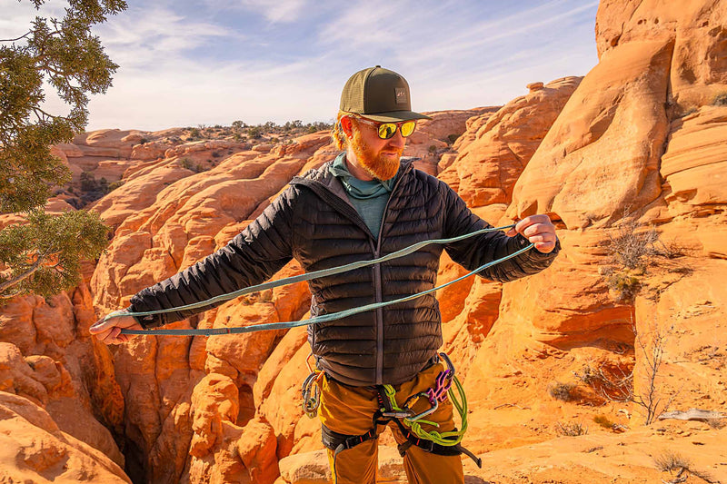Man wearing a Hunter Green colored Rover premium performance fitted hat from Hatchet Headwear, blue jacket, tan pants, and outdoor gear with climbing equipment in a desert landscape with red rock canyons in the background.