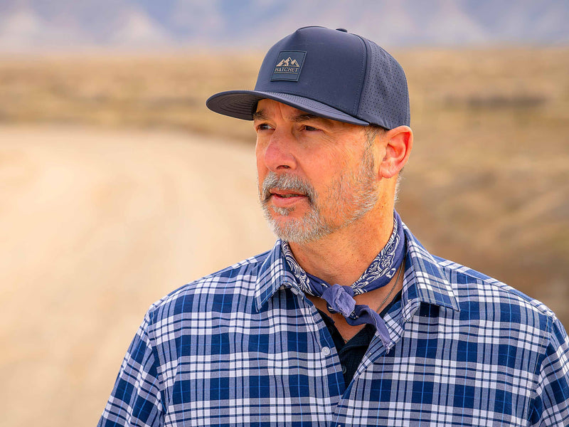 Man wearing a Slate Blue colored Rover premium performance fitted hat from Hatchet Headwear, a blue and white plaid shirt, and a blue and white handkerchief tied around his neck. The man is looking off into the distance while standing in front of a  blurred desert landscape.