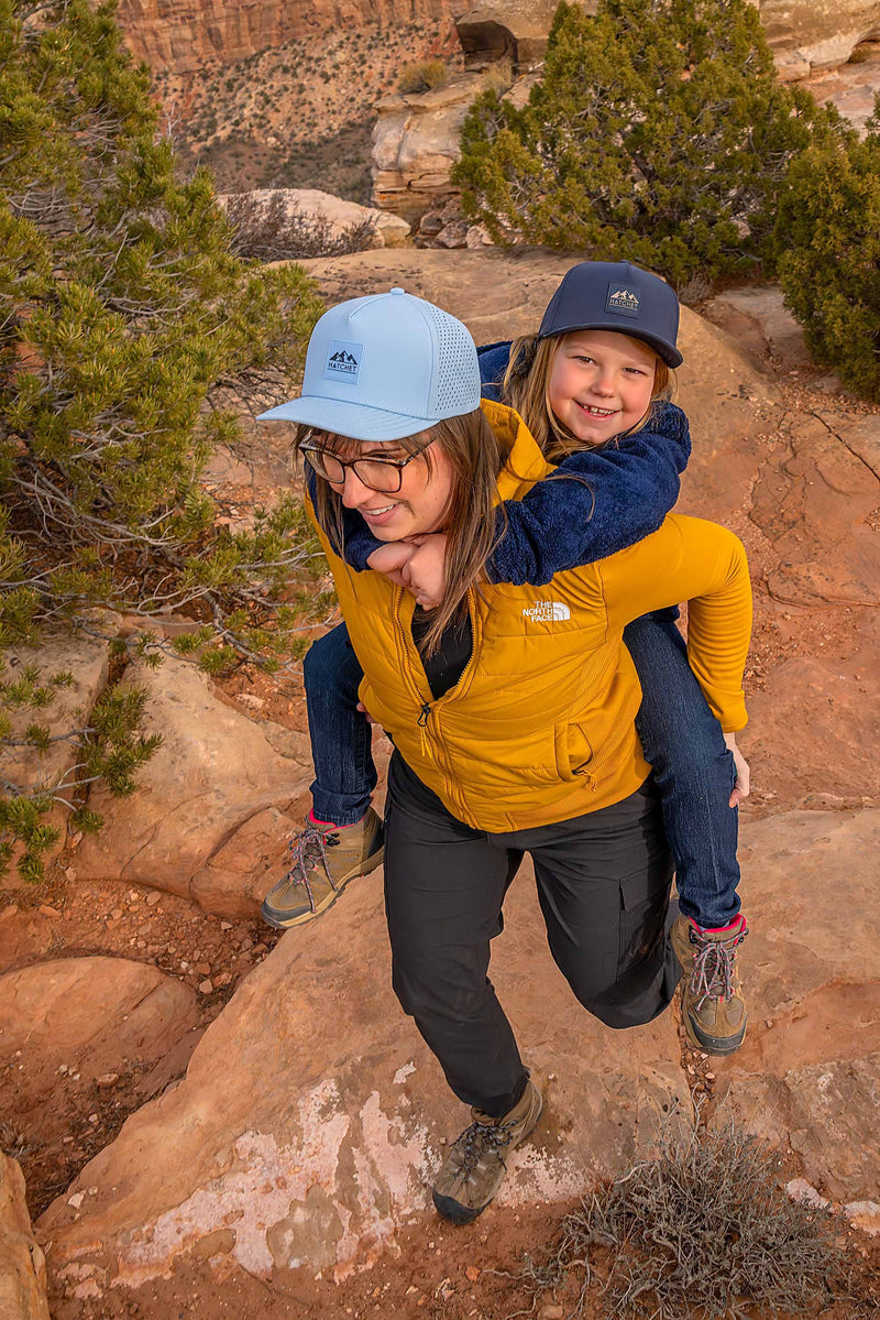 Mother and daughter wearing Rover premium performance fitted hats from Hatchet Headwear. Mother is wearing a Glacier colored Rover hat, a mustard yellow jacket, and charcoal hiking pants while carrying her daughter on her back during  a hike through a desert landscape.  Daughter is looking at the camera and wearing a Slate Blue colored Rover hat, a blue fleece jacket, and blue jeans.