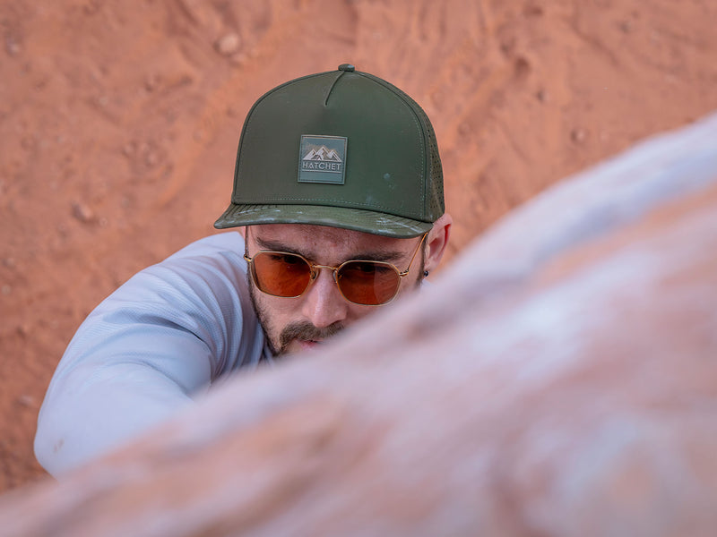 Man wearing a Hunter Green colored Rover premium performance fitted hat from Hatchet Headwear, sunglasses, and a gray long-sleeve outdoor shirt. The man is climbing a blurred red rock boulder with the desert ground's red dirt visible beneath him.