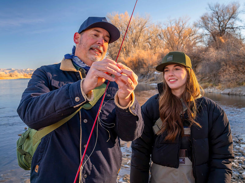 Father and daughter wearing Rover premium performance fitted hats from Hatchet Headwear. Father is wearing a Slate Blue colored Rover hat, blue jacket, and army green cross-body fishing bag while holding a fishing rod. Daughter is looking at the fishing rod that her dad is holding while wearing a Hunter Green colored Rover hat, black puffer jacket, and tan fly fishing bibs. The father and daughter are standing on the shore of a high desert lake preparing to fly fish.
