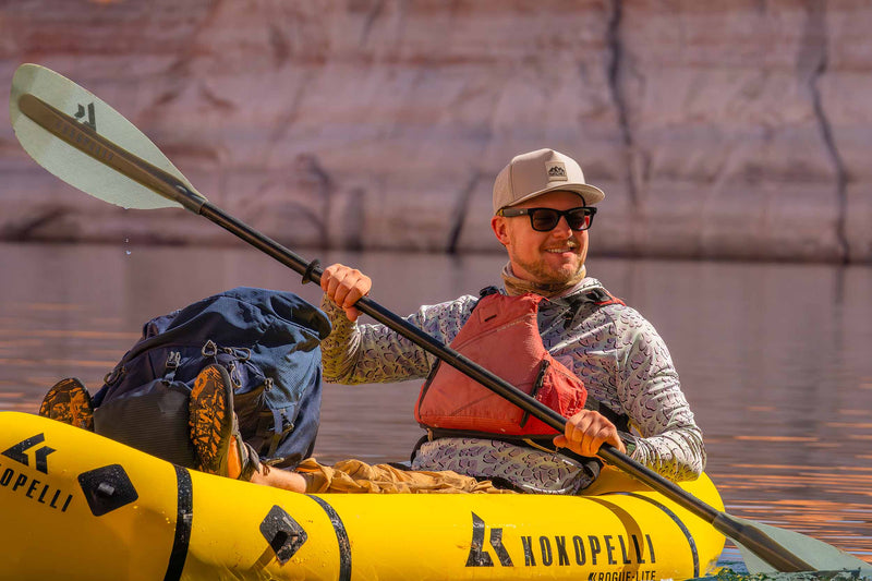 Man wearing a Boulder colored Rover premium performance fitted hat from Hatchet Headwear, a patterned long sleeve shirt, and an orange life vest while kayaking in a red rock canyon.  Man is smiling and facing towards the camera while paddling.