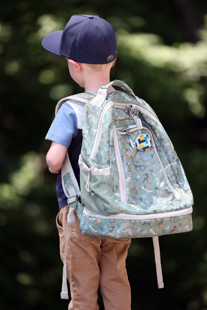 A child wearing a Slate Blue colored A woman wearing a Hunter Green colored Rover premium performance fitted hat from Hatchet Headwear, a light blue and navy blue t-shirt, and tan pants. The child is shown from behind while wearing a light green patterned backpack outdoors against a blurred green background.