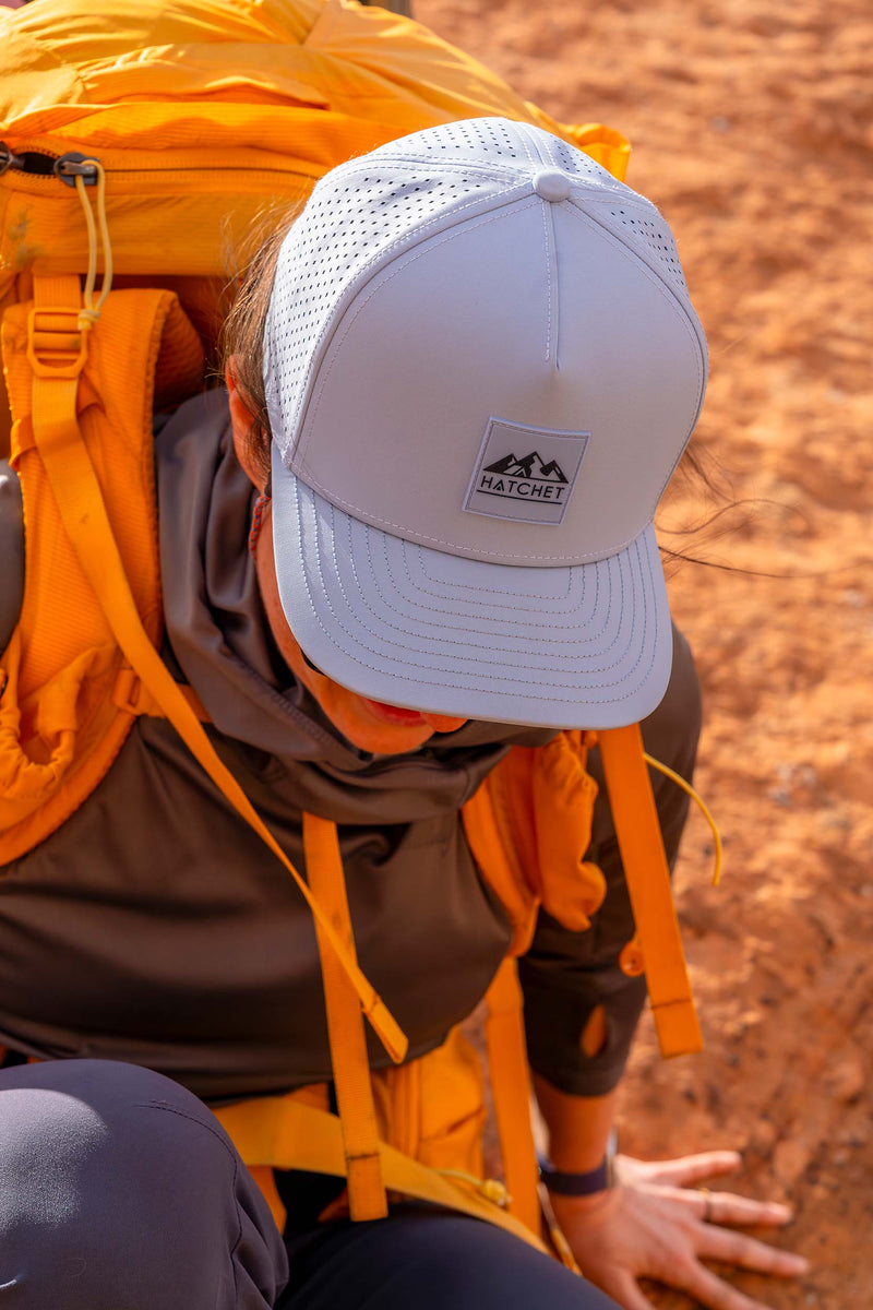 A close-up lifestyle shot of a hiker wearing a Glacier colored Rover premium performance fitted hat from Hatchet Headwear, crouching in a desert setting with an orange hiking backpack on their back.
